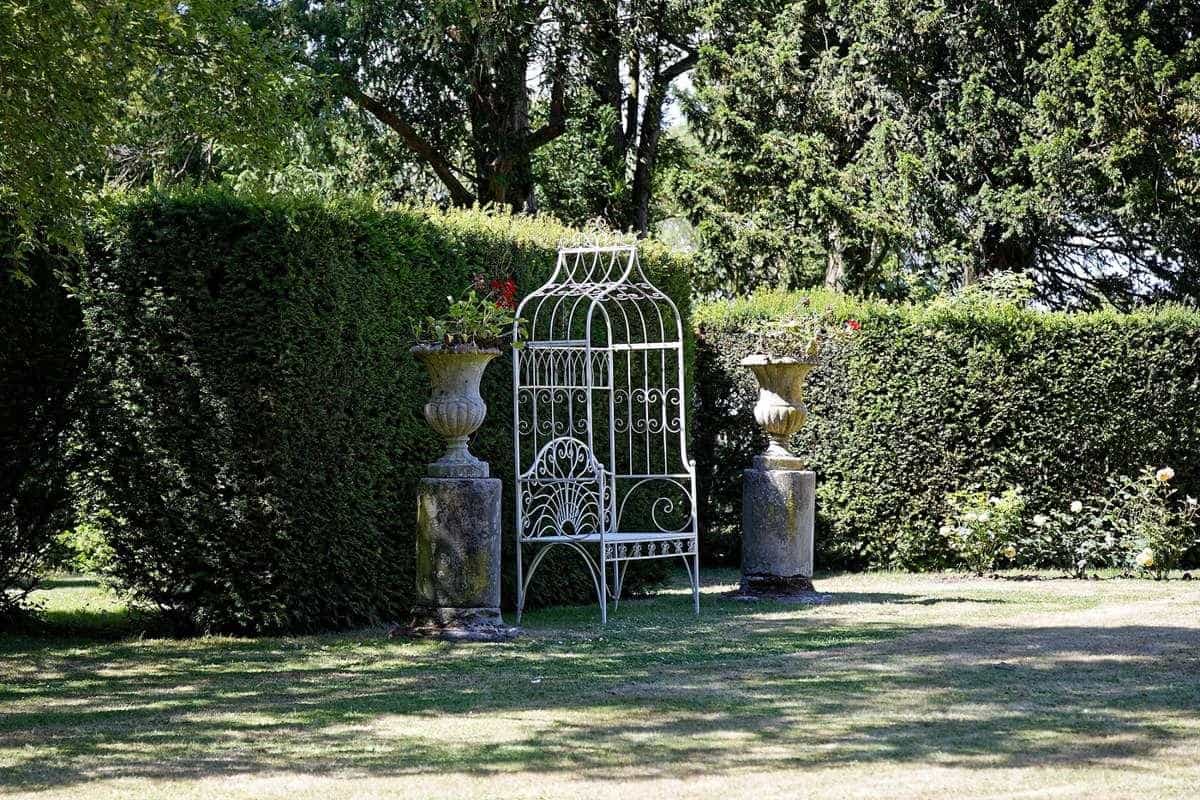 A white wrought-iron garden bench beneath an arbour surrounded by hedges