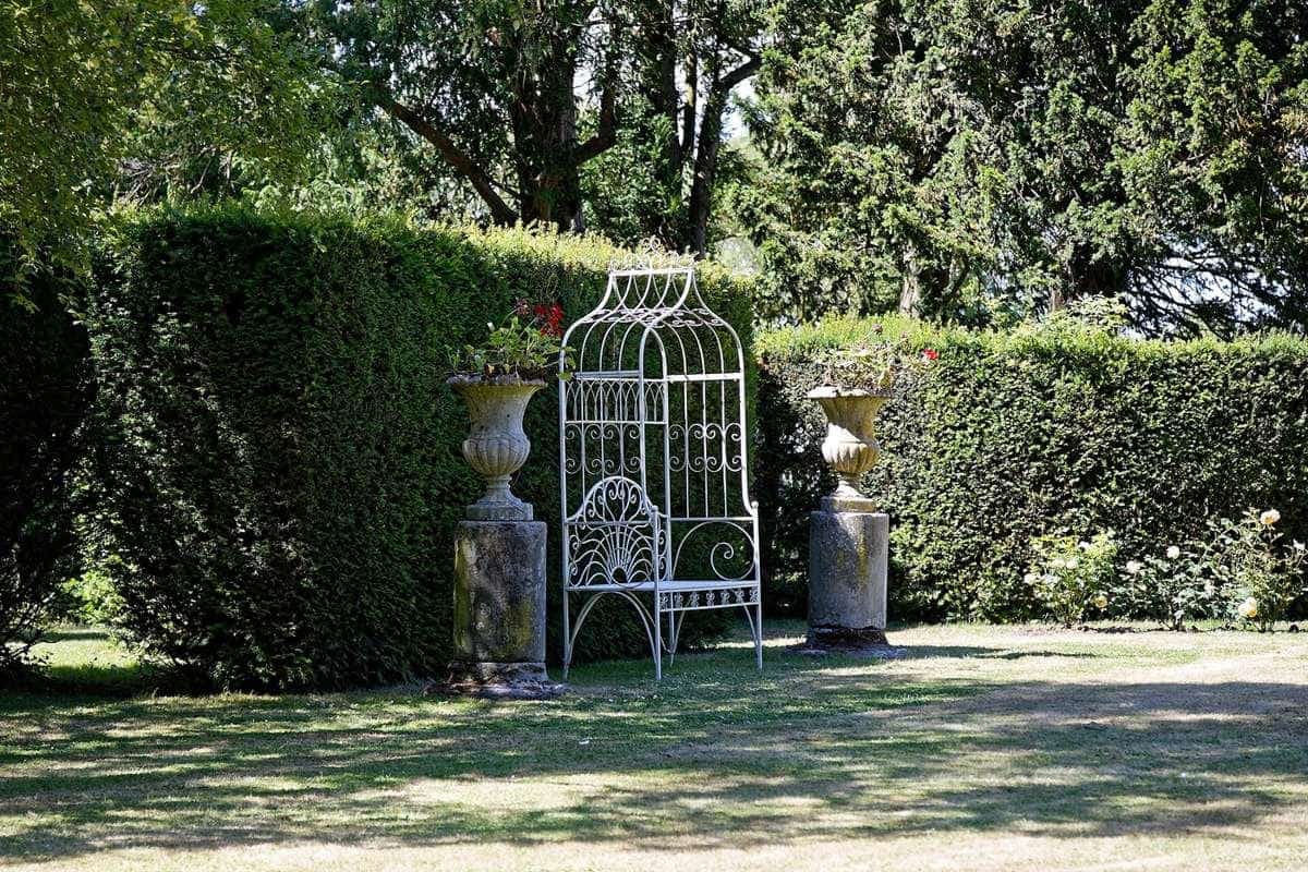 A white wrought-iron garden bench beneath an arbour surrounded by hedges